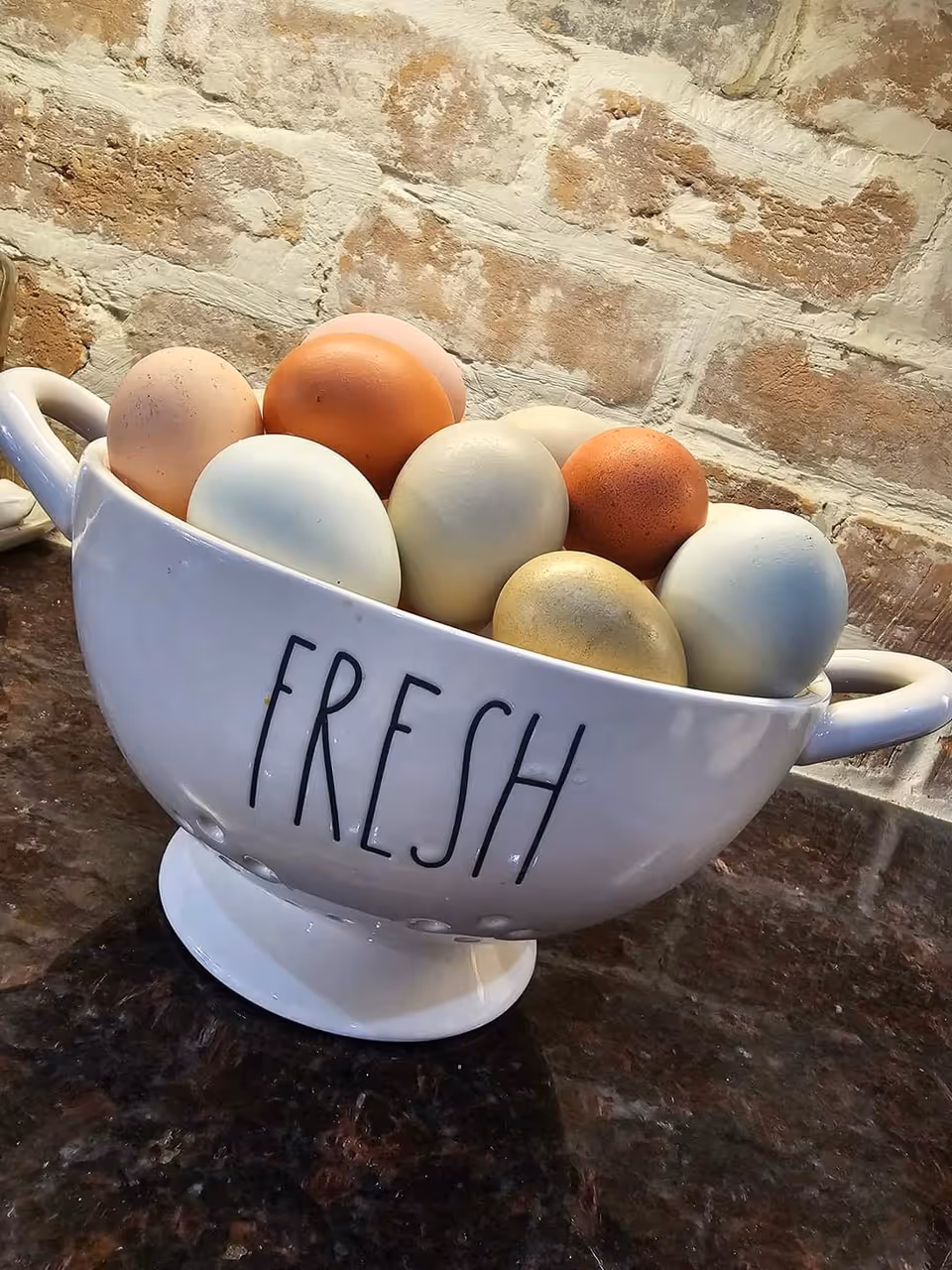 A white ceramic bowl with handles on both sides and the word 'FRESH' written on it, filled with a variety of brown, white, and light blue eggs, placed on a dark countertop with a brick wall background.