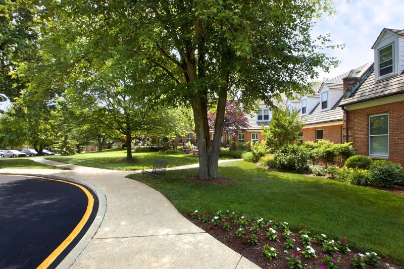 A sunny outdoor scene at Oak Bridge Terrace At Country House featuring a curved sidewalk bordered by a freshly paved road with a yellow line. There are green lawns, flower beds with blooming flowers, and several trees providing shade. A brick building with white dormer windows is visible on the right side, and a bench is placed near a tree along the sidewalk.