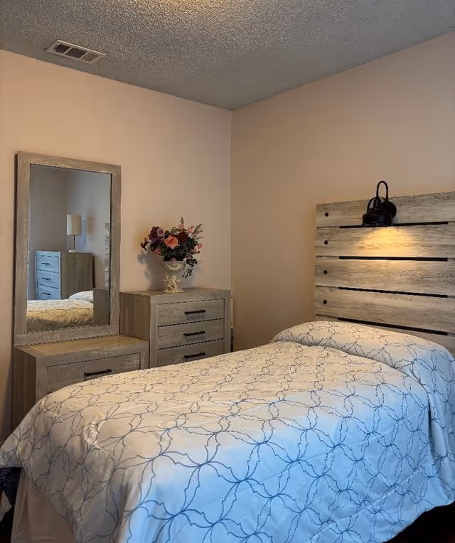 A small bedroom with a single bed covered in a patterned quilt, a rustic wood headboard with a lamp, and a matching dresser and mirror topped with a vase of flowers.