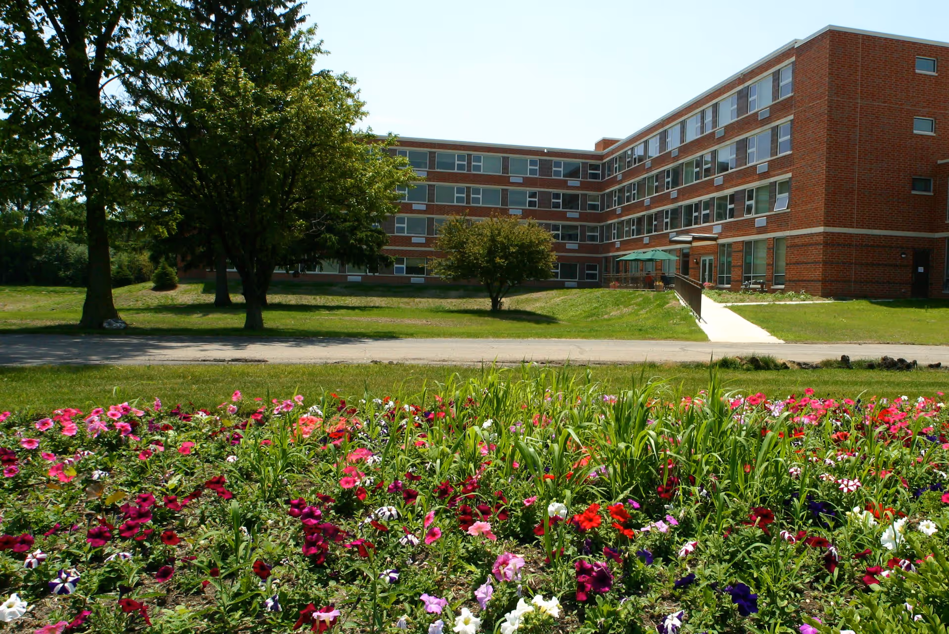 A multi-story brick building with many windows, surrounded by green grass and trees. In the foreground, there is a colorful flower bed with various blooming flowers. A paved walkway leads to the building entrance, which has a green umbrella and outdoor seating.