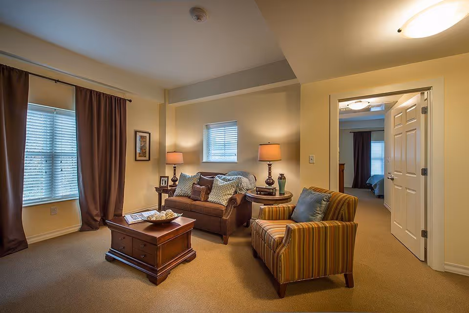A cozy living room with a brown loveseat adorned with multiple pillows, a striped armchair with a blue cushion, a wooden coffee table with decorative items, two side tables with lamps, and windows with brown curtains. An open door reveals a bedroom in the background.