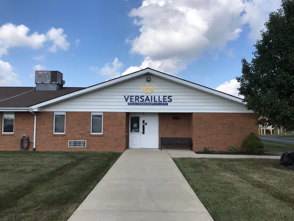 Front exterior view of Versailles Rehabilitation and Healthcare Center building with a brick facade, white siding on the upper portion, a concrete walkway leading to a white door entrance, a bench to the right of the door, and a tree on the right side under a partly cloudy sky.