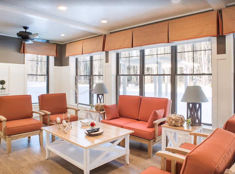 Bright common lounge with coral-upholstered chairs and sofas around a wooden coffee table in front of large windows.