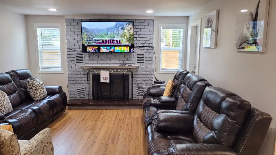 A cozy living room with two dark brown leather sofas facing each other across a wooden floor. A flat-screen TV is mounted above a white-painted brick fireplace with a black screen. Two windows with white blinds flank the fireplace, and there are decorative pillows on the sofas. The walls are light-colored with framed artwork.