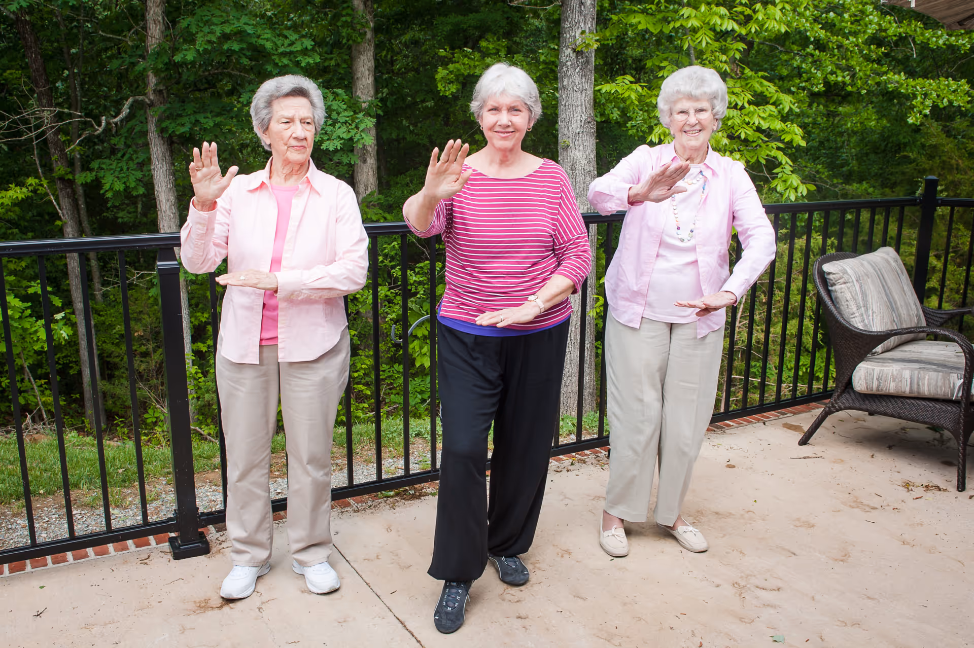 Three elderly women standing outdoors on a patio with a black metal railing and green trees in the background, performing a synchronized exercise or Tai Chi with one hand raised and the other hand held horizontally in front of them.