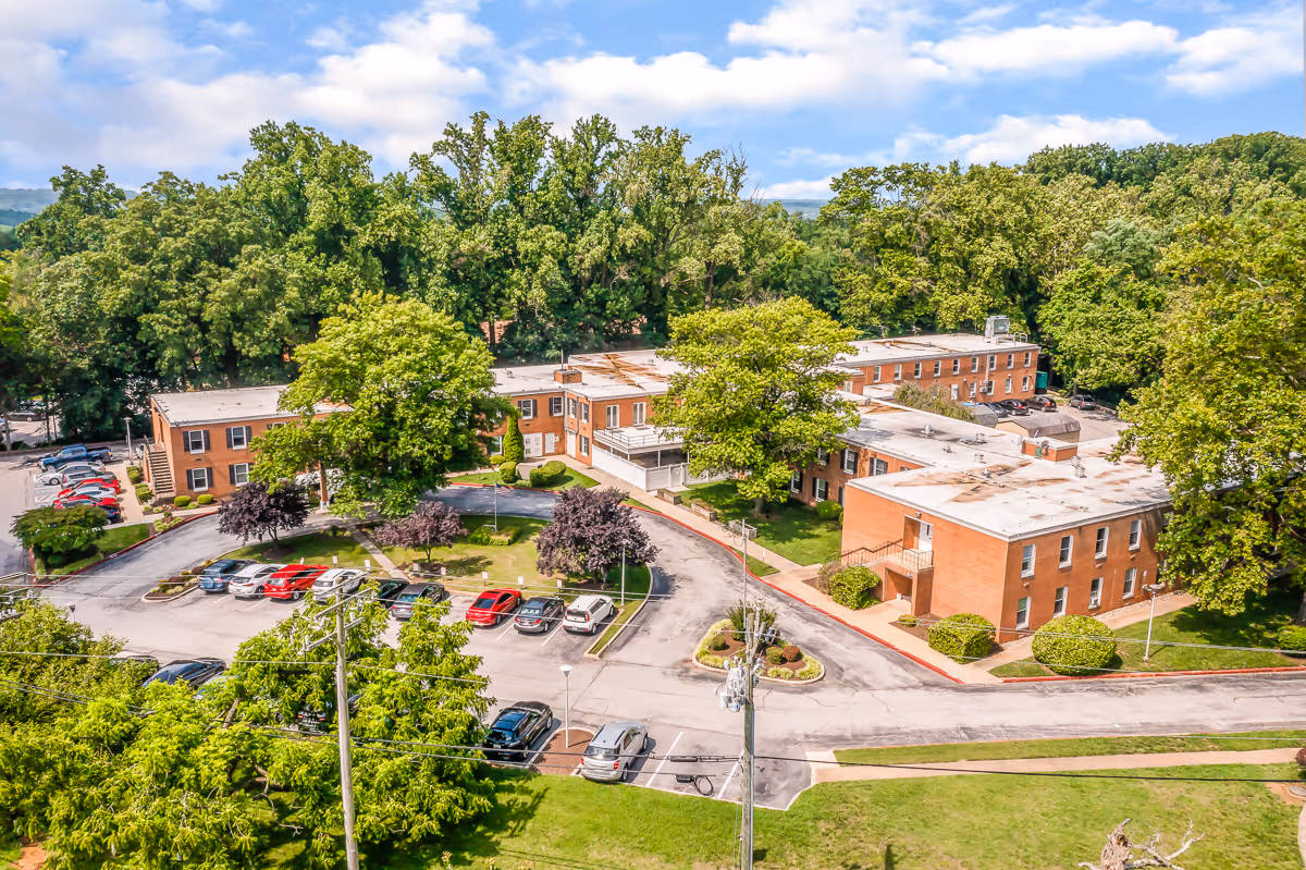 Aerial view of Autumn Lake Healthcare at Summit Park showing a large, U-shaped brick building surrounded by trees and greenery. There is a parking lot with several cars parked, and the area is well-maintained with landscaped bushes and grass. The sky is partly cloudy.