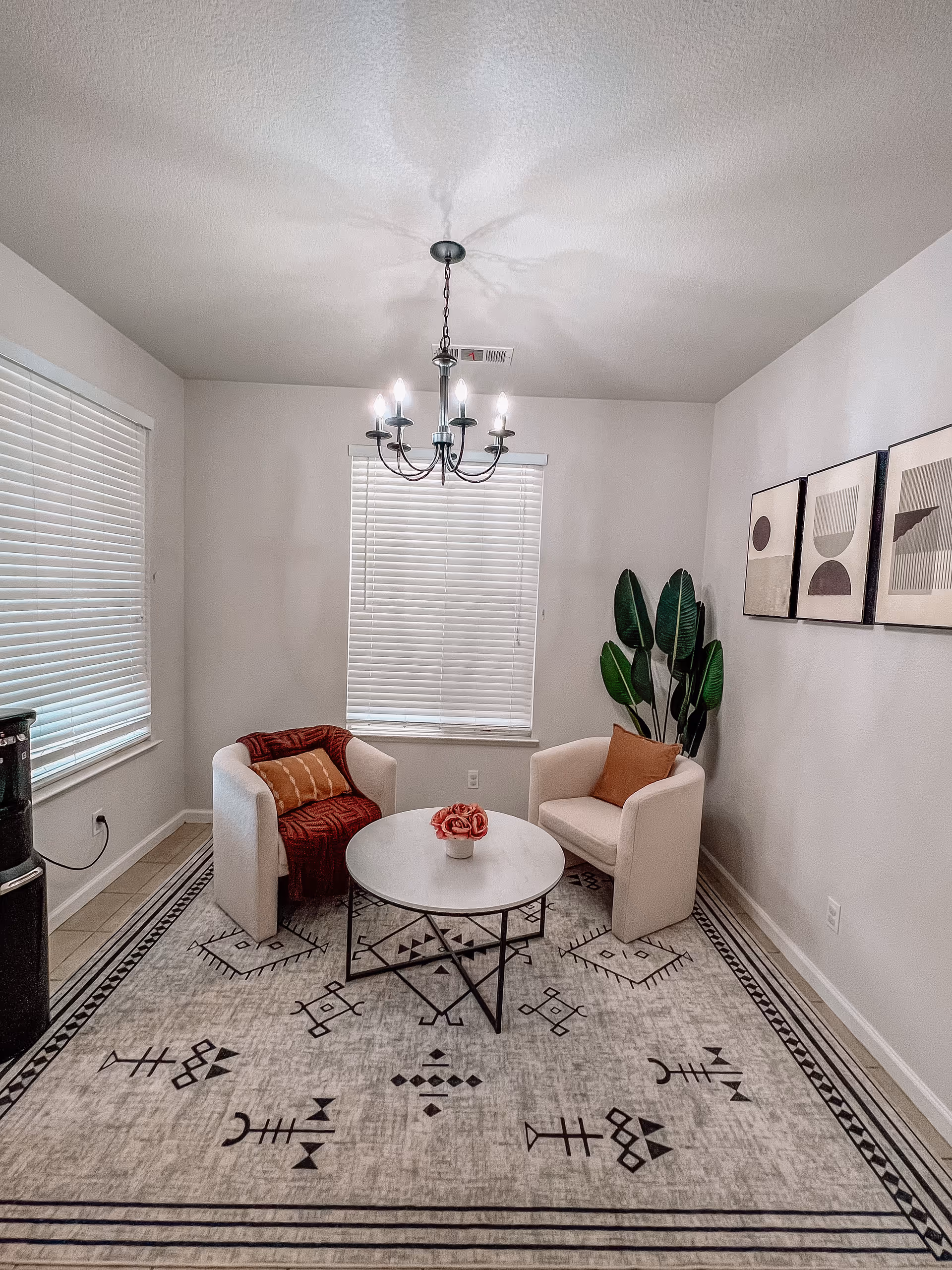 Cozy sitting area with two upholstered armchairs around a round coffee table on a patterned rug, a hanging chandelier, potted plant, and wall art.