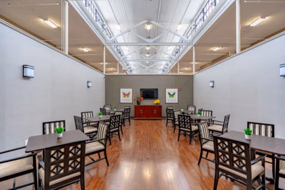 A bright dining area with wooden floors, several tables and chairs arranged neatly, small green plants on each table, and a cabinet with a TV and butterfly artwork on the far wall under a high ceiling with skylights.