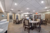 A spacious common area with multiple round tables and chairs arranged for seating. The room has a neutral color scheme with beige walls and carpeted floors. There are windows with curtains, framed artwork on the walls, and a cabinet with glass doors. The ceiling has recessed lighting, and an exit sign is visible above a door.