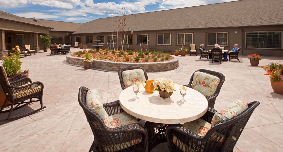 Outdoor courtyard area at Spring Creek Inn Memory Care Community featuring a round table with four cushioned wicker chairs, a flower centerpiece, and glasses on the table. In the background, there are additional seating areas with people sitting and a raised garden bed with plants. The building surrounds the courtyard under a partly cloudy sky.