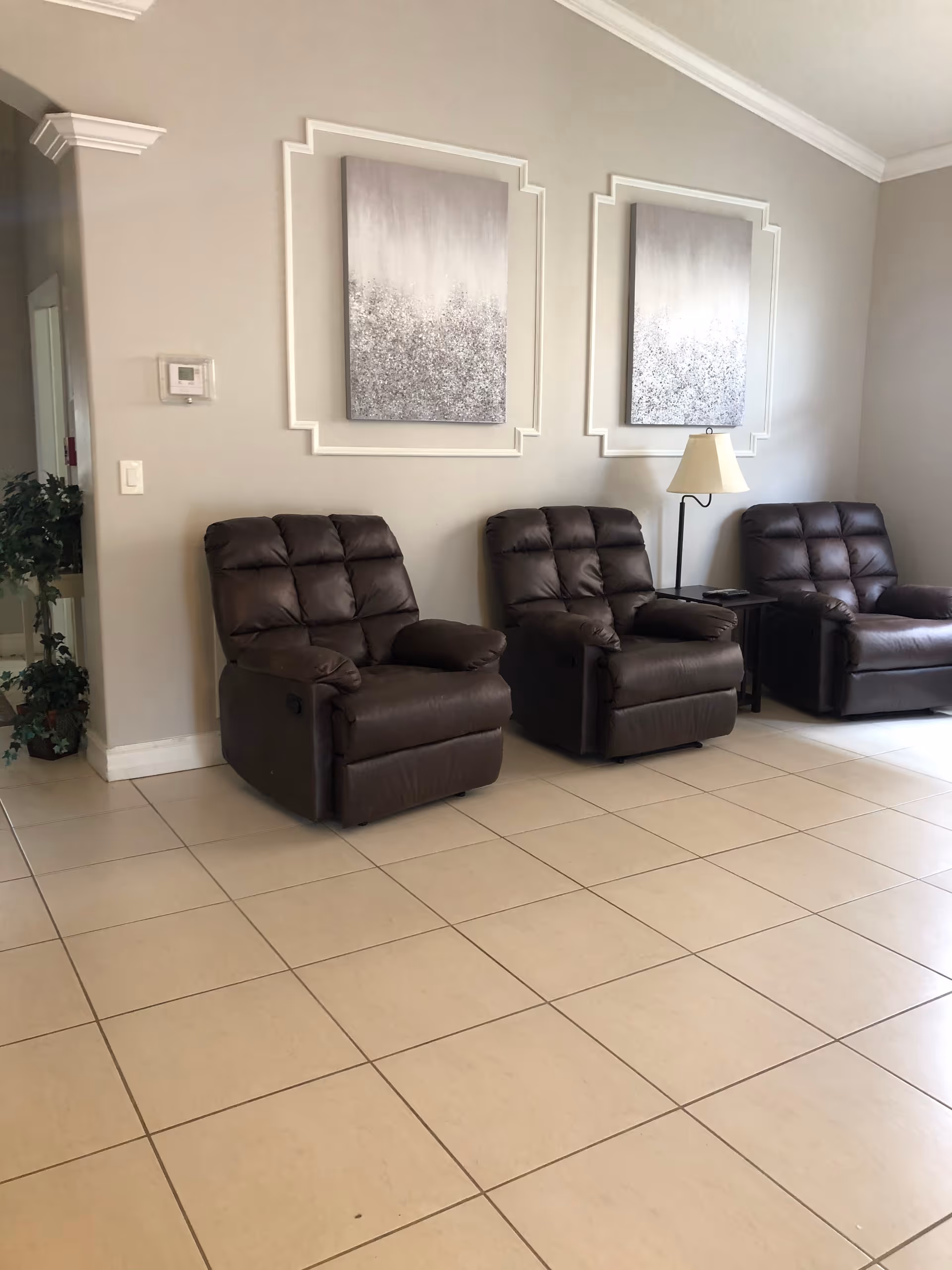A room with three dark brown leather recliner chairs arranged side by side against a light beige wall. Above the chairs are two abstract paintings with gray and white tones, each framed with decorative molding on the wall. A small table with a lamp is placed between the second and third chairs. The floor is tiled with large beige tiles, and there is a small plant visible in the corner near a doorway.