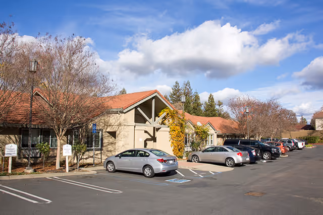 Exterior view of Brookdale Roseville facility showing a single-story building with a red-tiled roof, several parked cars in front, leafless trees, and a partly cloudy sky.