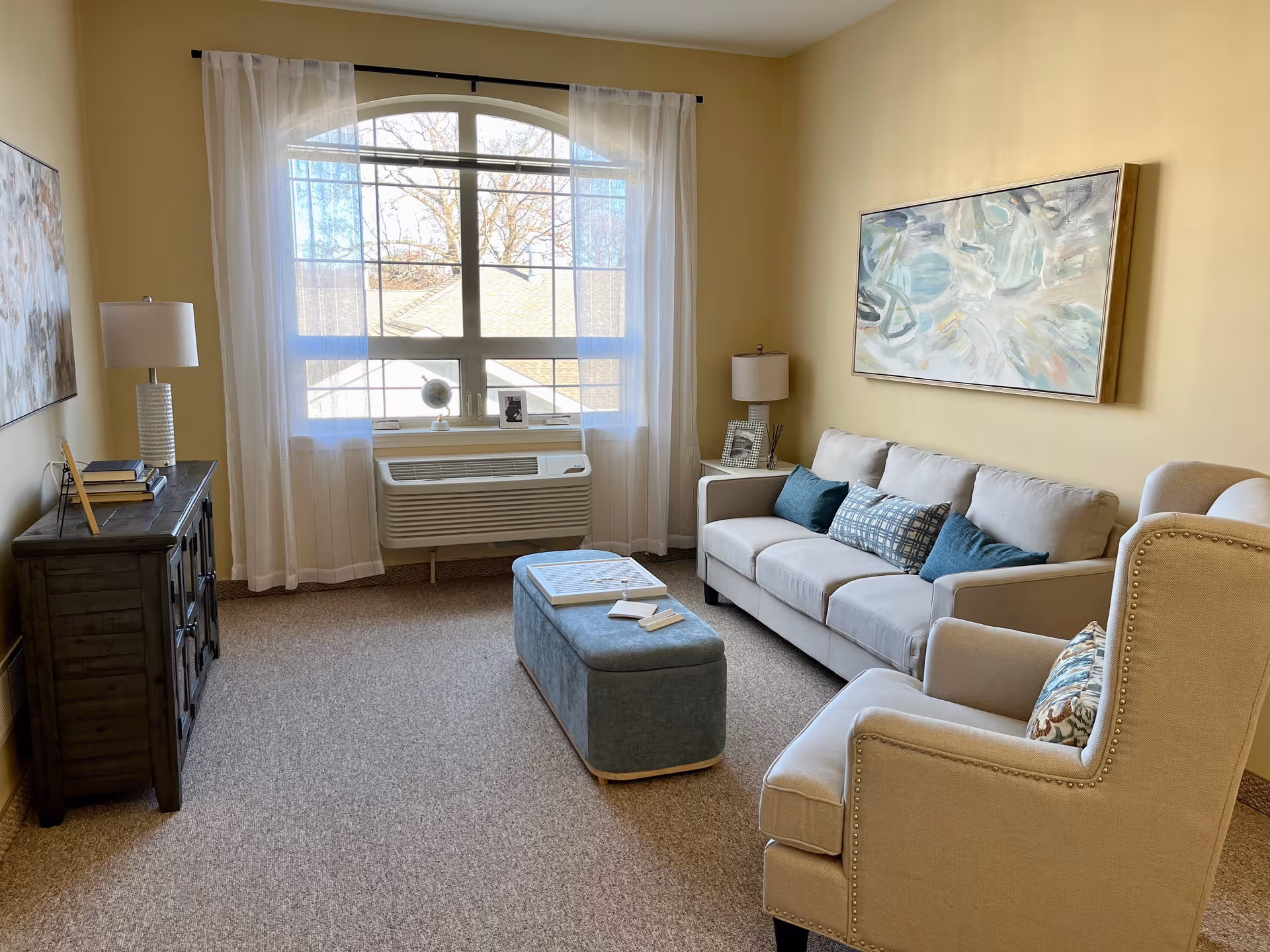 A cozy living room with a beige sofa adorned with blue and patterned cushions, a matching armchair with a decorative pillow, and a blue upholstered ottoman serving as a coffee table. The room features a large arched window with sheer white curtains, letting in natural light. On the left side, there is a dark wooden cabinet with books and a lamp, and another lamp is on a small side table next to the sofa. Abstract paintings hang on the walls, and the carpet is a neutral tone.