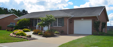 Single-story brick ranch-style building with an attached garage, front porch, walkway, and landscaped lawn under a partly cloudy sky.