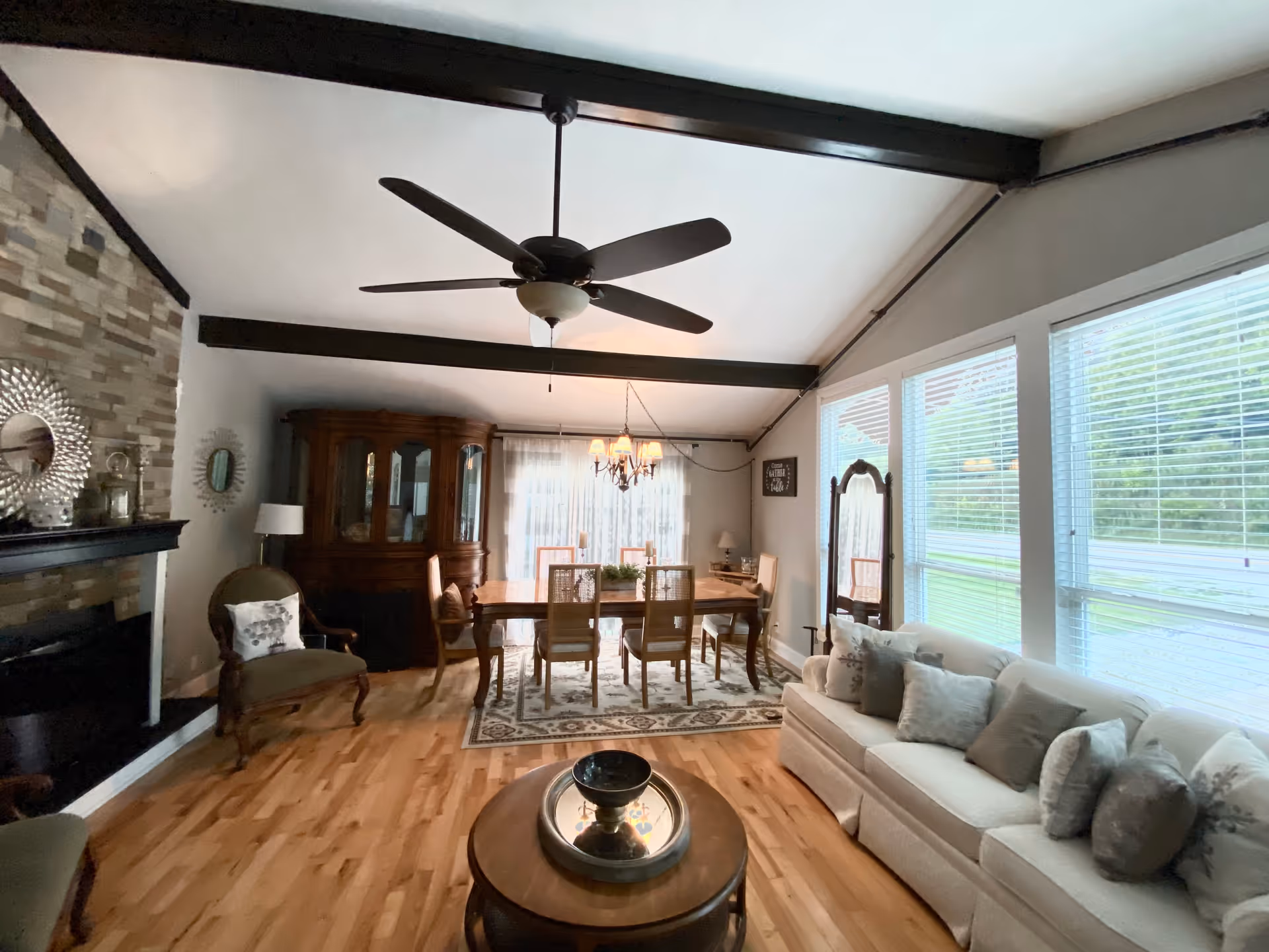 Bright open living and dining area with vaulted beamed ceiling, ceiling fan, large windows, sofa, coffee table, dining table and fireplace.