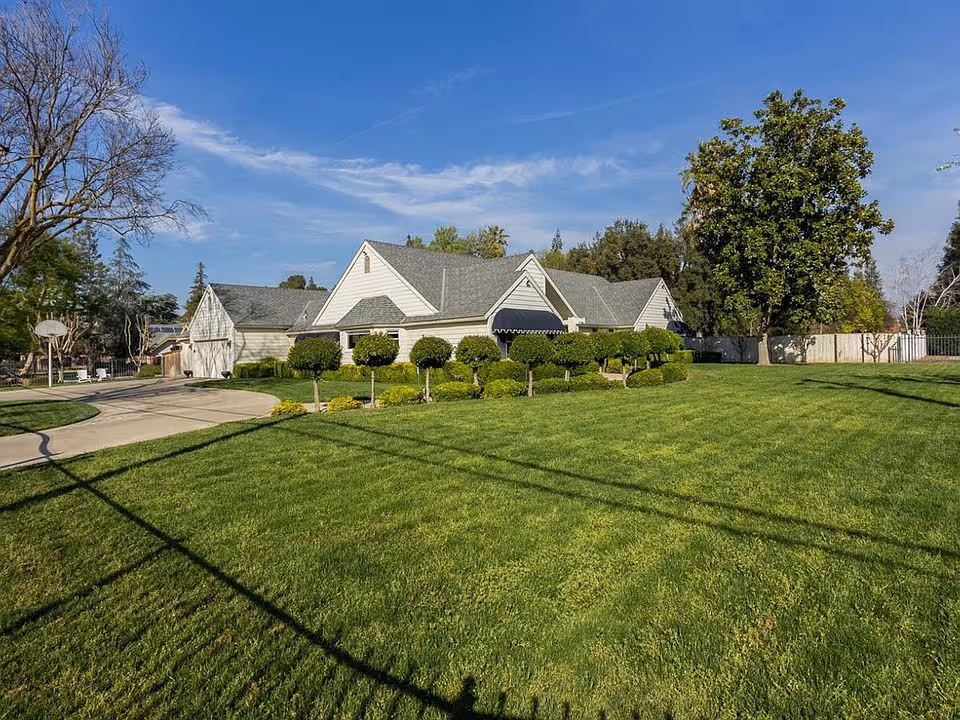 Single-story white house with a large manicured front lawn, trimmed shrubs, driveway and trees under a blue sky.