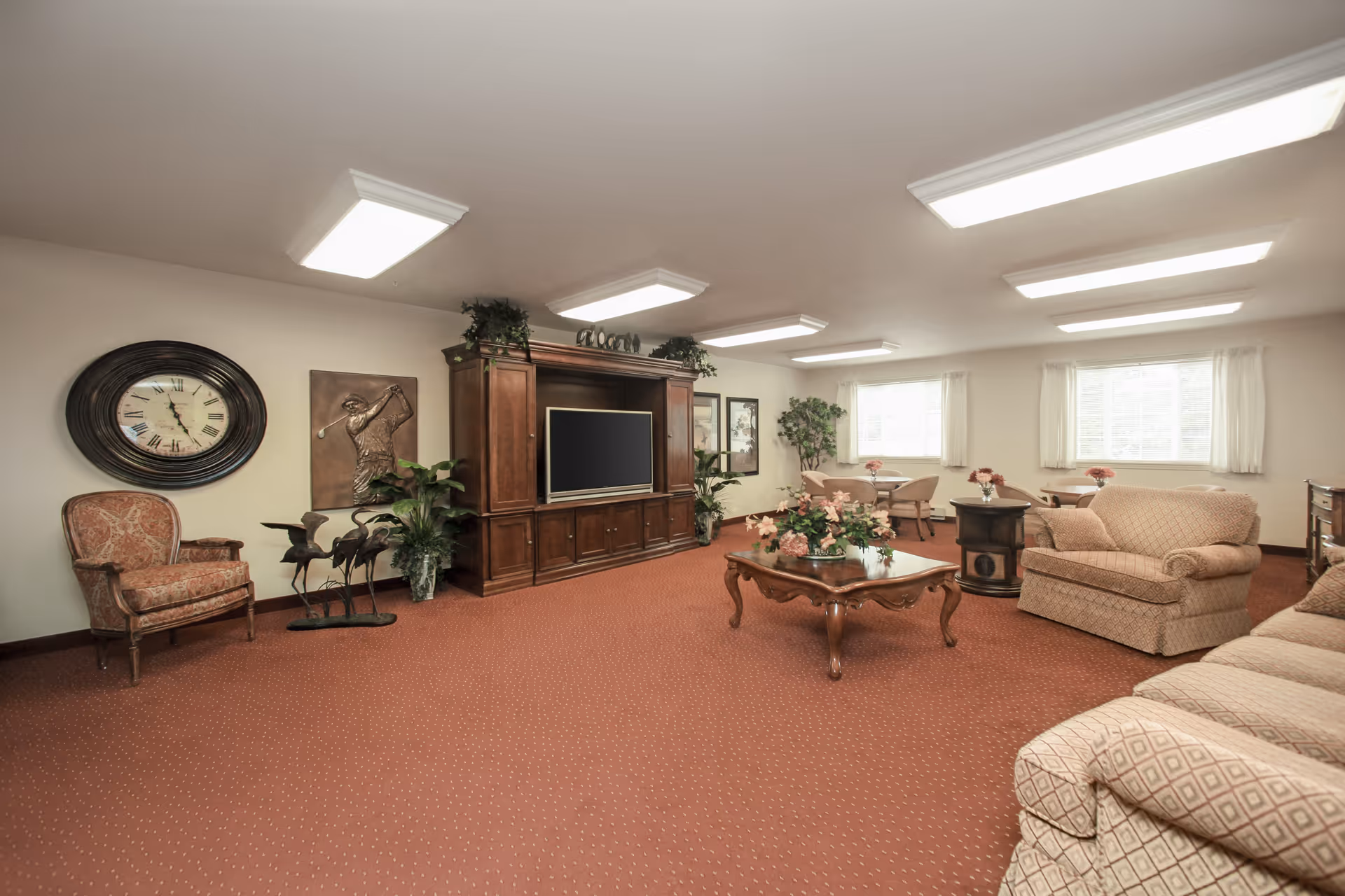A spacious living room with beige walls and carpeted floor in a warm reddish tone. The room features a large wooden entertainment center with a flat-screen TV, several upholstered armchairs and sofas in beige tones, a wooden coffee table with floral arrangements, and decorative plants. There is a large wall clock, a golf-themed wall art, and multiple windows with white curtains allowing natural light to enter.