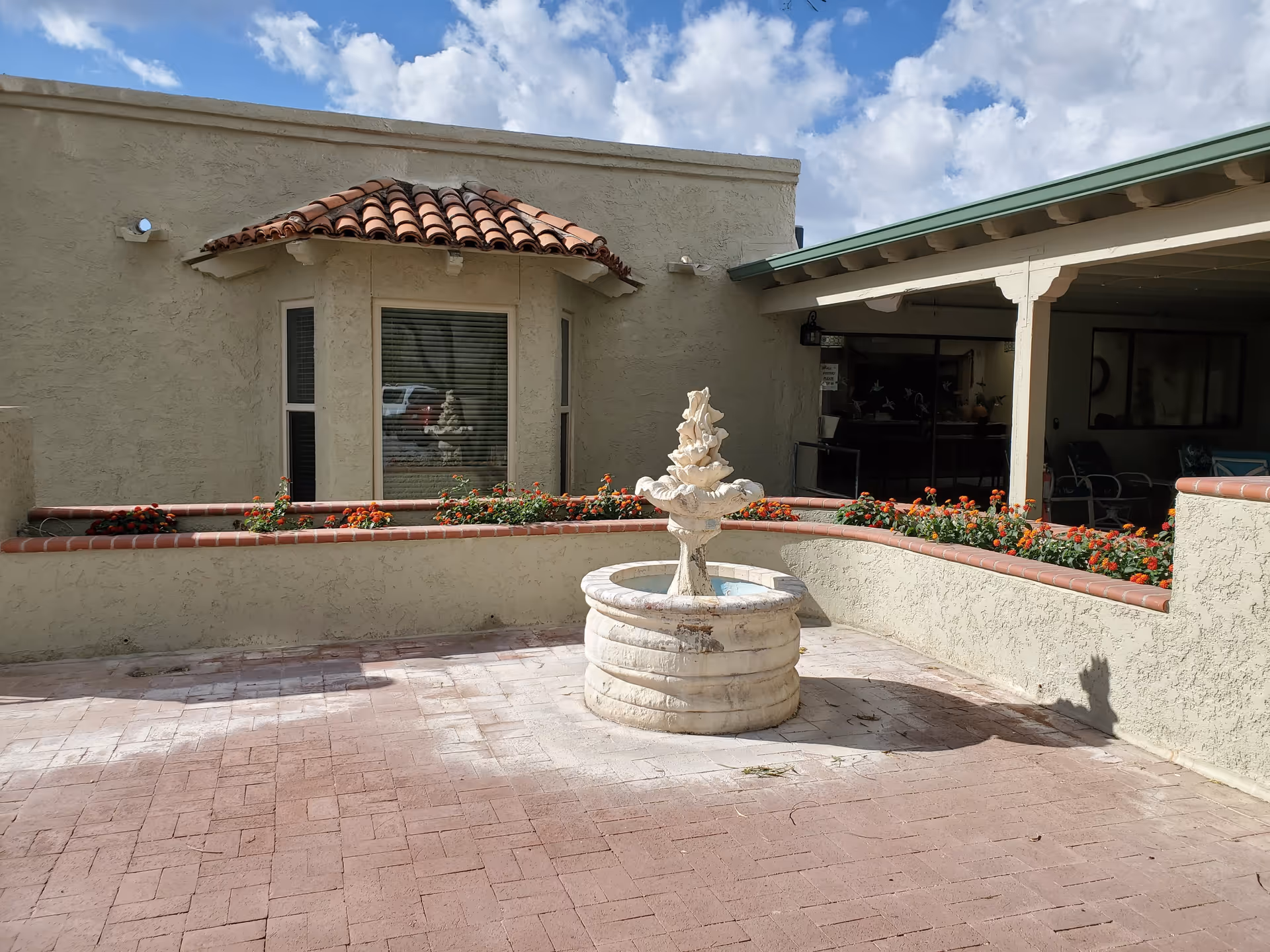 Courtyard featuring a central stone fountain, planter beds with red flowers, and a stucco building with a covered patio under a partly cloudy sky.
