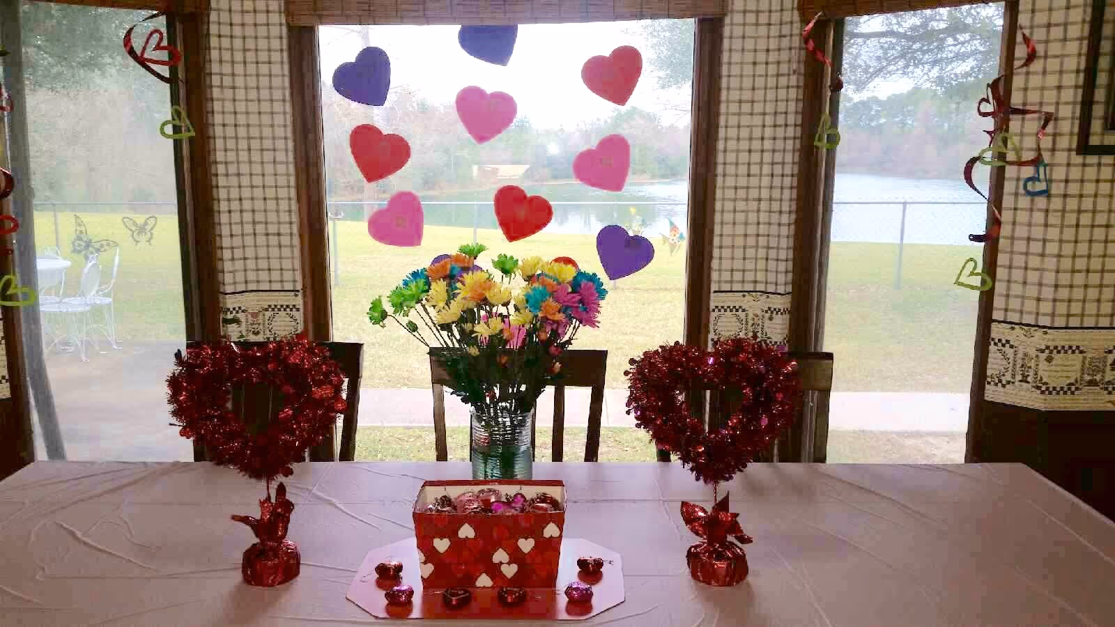 A dining table decorated with Valentine's Day themed items including two red tinsel heart-shaped decorations, a red box with white hearts filled with chocolates, and a vase of colorful flowers. The background shows large windows with heart-shaped paper cutouts and a view of a grassy outdoor area with a lake.