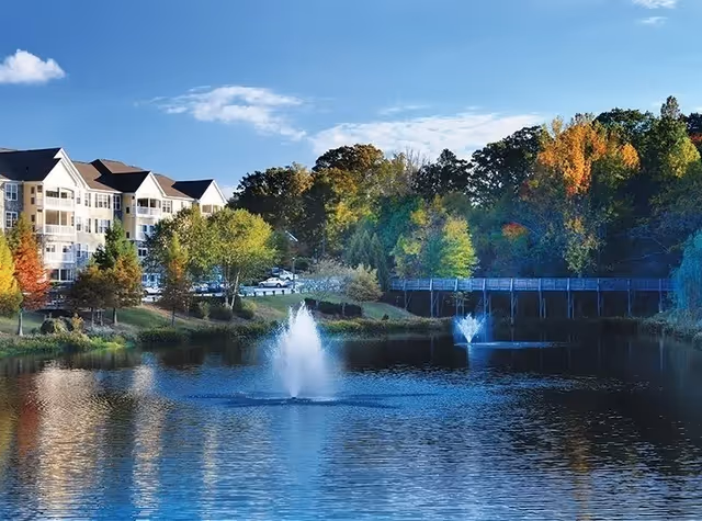 A serene outdoor scene featuring a large pond with two water fountains, surrounded by trees with autumn foliage. In the background, there is a multi-story residential building and a wooden walkway or bridge extending through the trees.