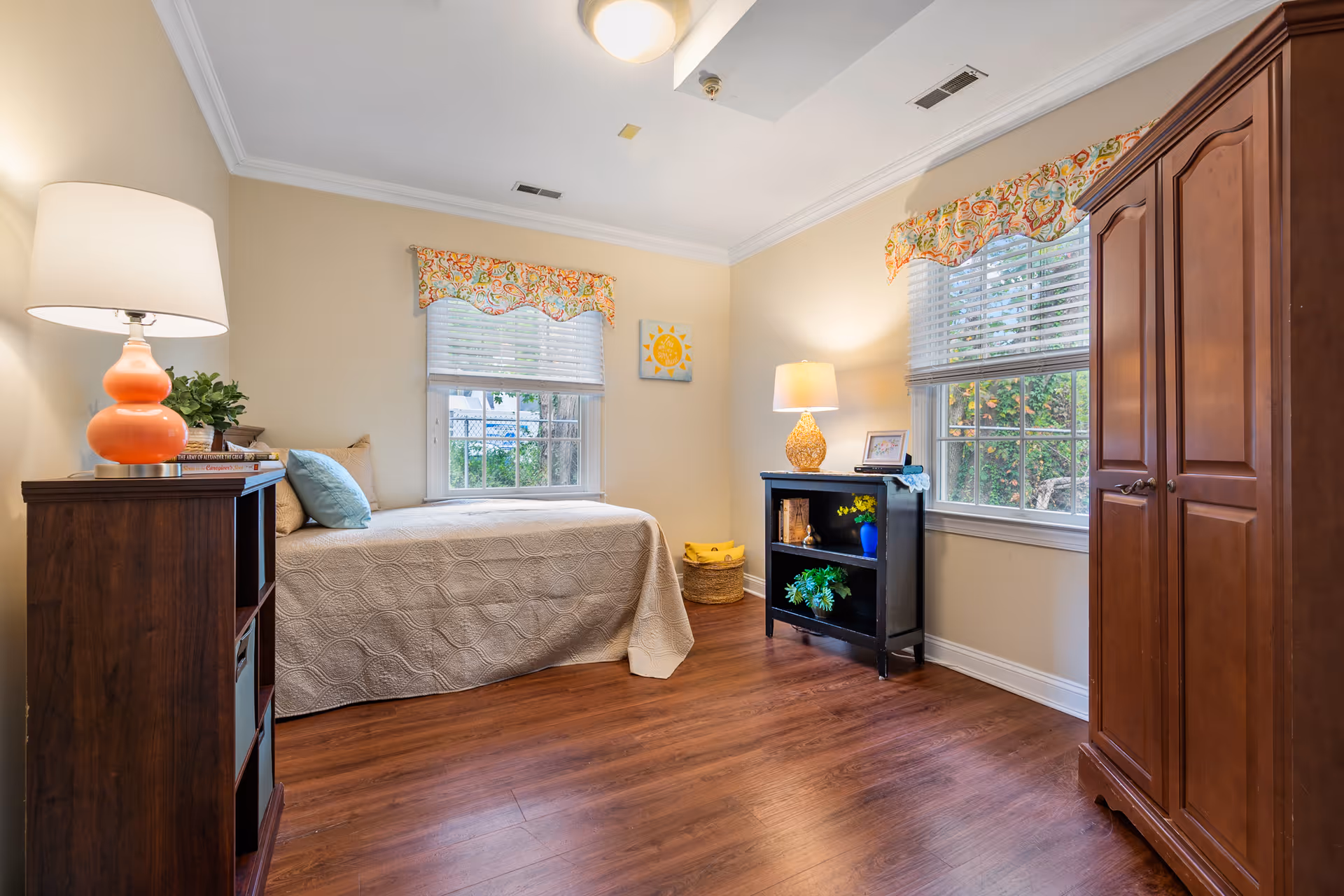 A cozy bedroom with a single bed covered in a beige quilt, two windows with colorful valances, a wooden wardrobe, a small black bookshelf with a lamp and decorative items, and a wooden floor.