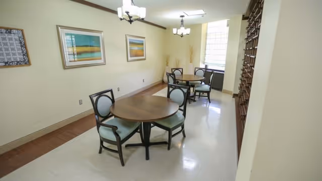 A small dining area with two round wooden tables, each surrounded by four chairs with light blue cushions. The room has cream-colored walls adorned with two framed landscape paintings and a decorative wall hanging. There are two ceiling light fixtures and a large window letting in natural light. A wooden mail organizer is mounted on the right wall.