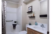 A clean and modern bathroom featuring a white bathtub with a patterned shower curtain, a built-in grab bar, a small potted plant on the bathtub ledge, a white toilet, a wooden vanity with a white countertop, a silver faucet, a soap dispenser, a small decorative item, a mirror, and two wooden floating shelves with framed artwork and a small plant.