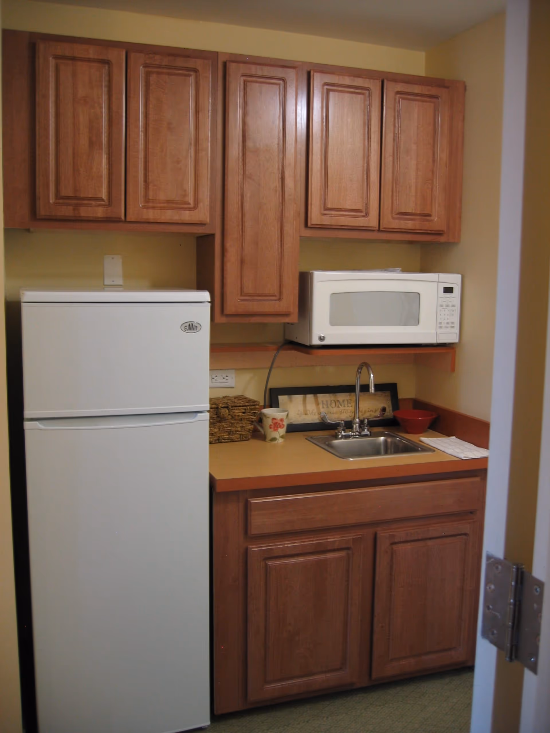 Small kitchen area with wooden cabinets, a white refrigerator, a white microwave on a shelf, a sink with a faucet, a cup, a red bowl, and a decorative framed sign on the countertop.