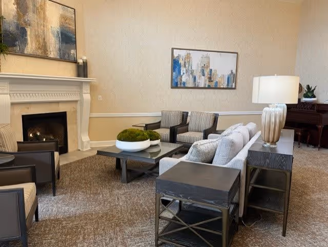 A cozy senior living room with a fireplace, a white mantel, and a fire burning inside. The room features a gray sofa, two striped armchairs, a dark wood coffee table with decorative green moss balls, a side table with a lamp, and abstract artwork on the walls. There is also a piano in the background.