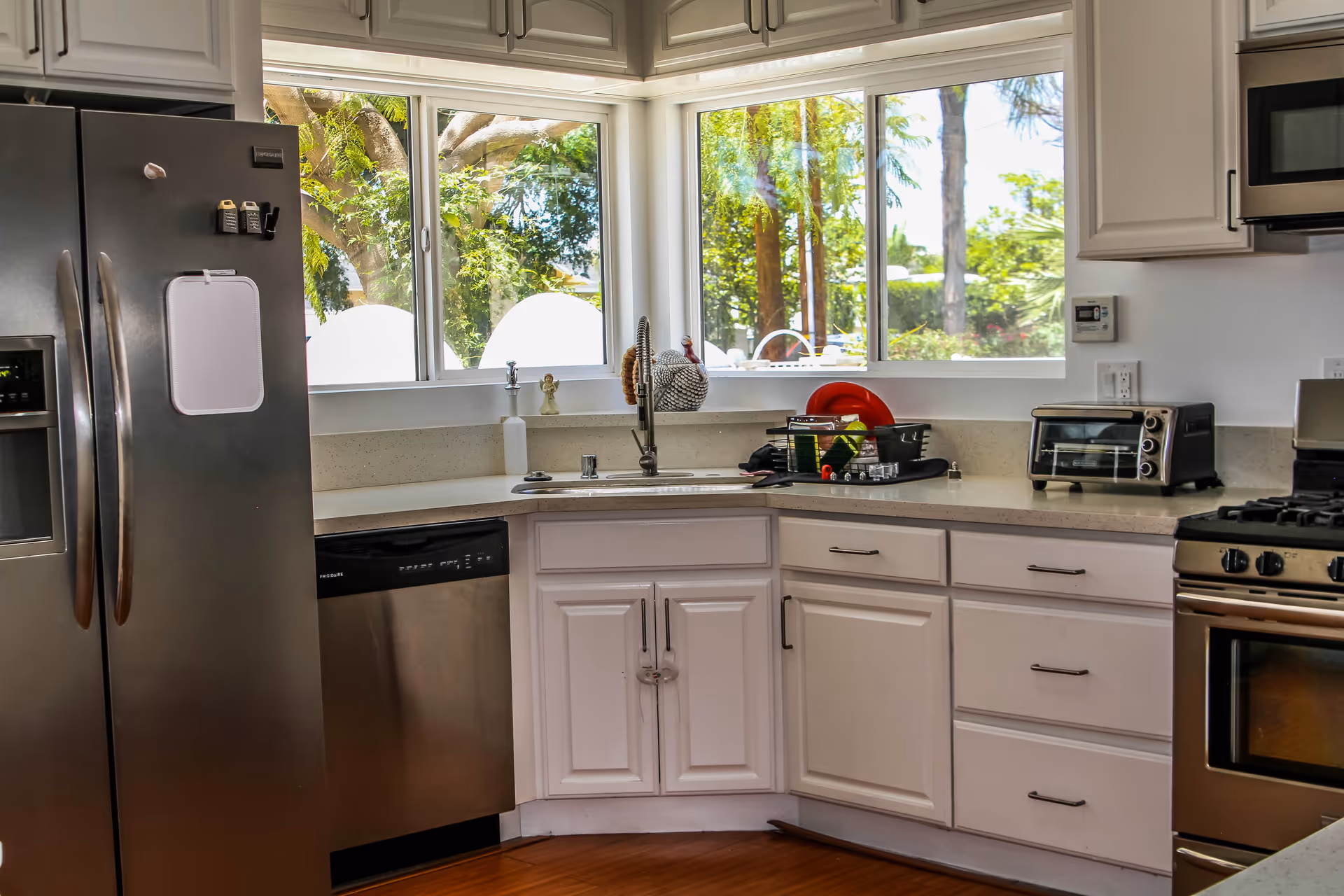 Bright kitchen with white cabinets, stainless steel refrigerator, dishwasher, oven, and microwave. A corner sink is positioned under large windows showing green trees outside. Countertop holds a dish rack with dishes and a small toaster oven.
