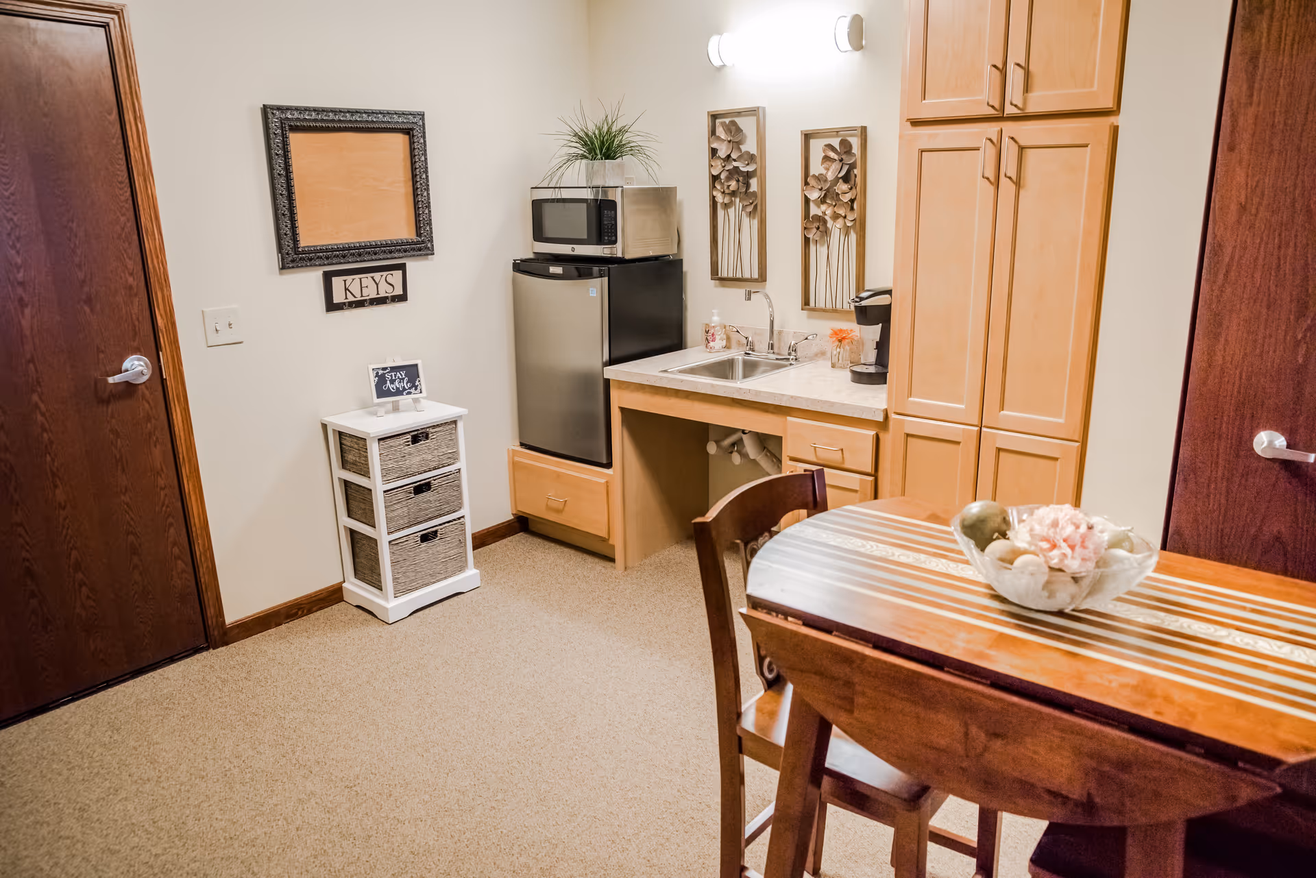 A small kitchenette area with a stainless steel mini fridge topped by a microwave, a sink with a coffee maker on the counter, wooden cabinets, and a wooden table with chairs. There is a small white storage unit with three drawers and a sign that says 'STAY Awhile' on top. The walls are decorated with a framed corkboard, a 'KEYS' sign, and two floral metal wall art pieces.