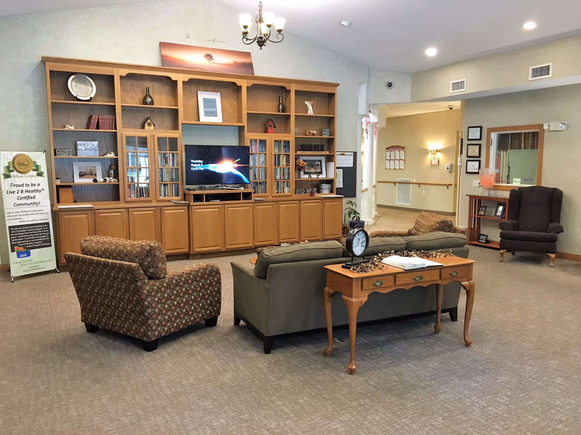 A cozy senior living facility common area with a large wooden entertainment center holding a flat-screen TV, books, and decorative items. In front of the entertainment center are a patterned armchair and a green sofa with a wooden side table holding a clock and some decorations. The room has beige carpet, soft lighting, and a hallway leading to other rooms. A banner on the left side promotes a wellness certification.