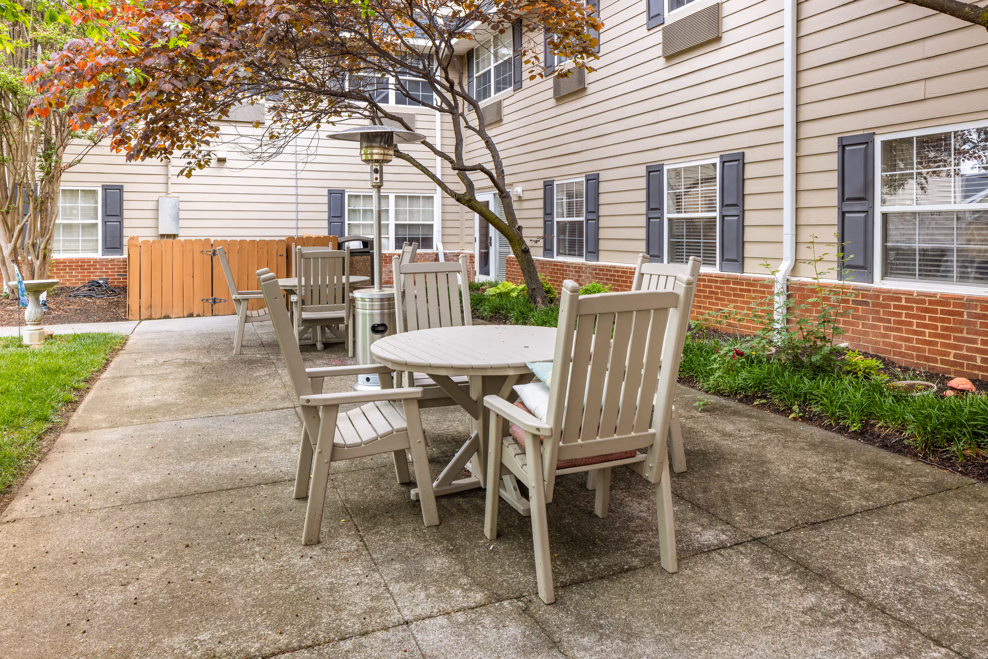 Outdoor patio area with beige wooden chairs and round tables on a concrete surface, surrounded by a garden bed with green plants and a tree with reddish leaves. The building exterior features beige siding with brick accents and multiple windows with black shutters.