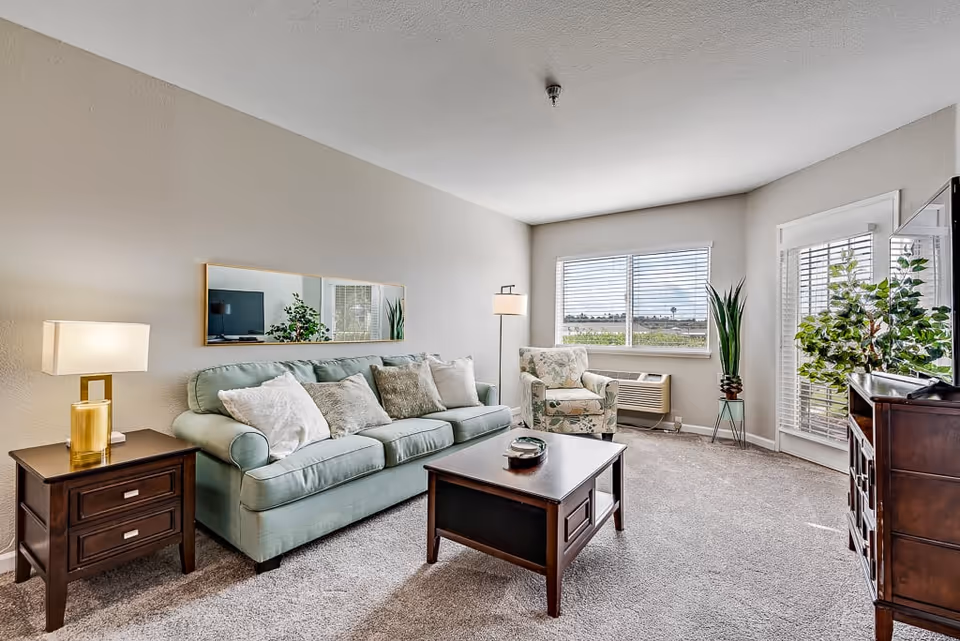 A bright and cozy living room with a light green sofa adorned with several cushions, a floral armchair near a window, a wooden coffee table, a side table with a lamp, a tall floor lamp, a large mirror on the wall, and a wooden cabinet with a TV and a potted plant. The room has beige walls, carpeted floor, and a window with blinds letting in natural light.