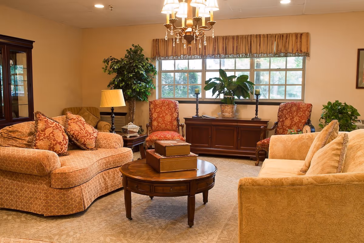 A cozy living room with two patterned sofas, two red upholstered armchairs, a round wooden coffee table with decorative boxes, a wooden cabinet under a large window with plants and candles, a floor lamp, and a chandelier hanging from the ceiling.