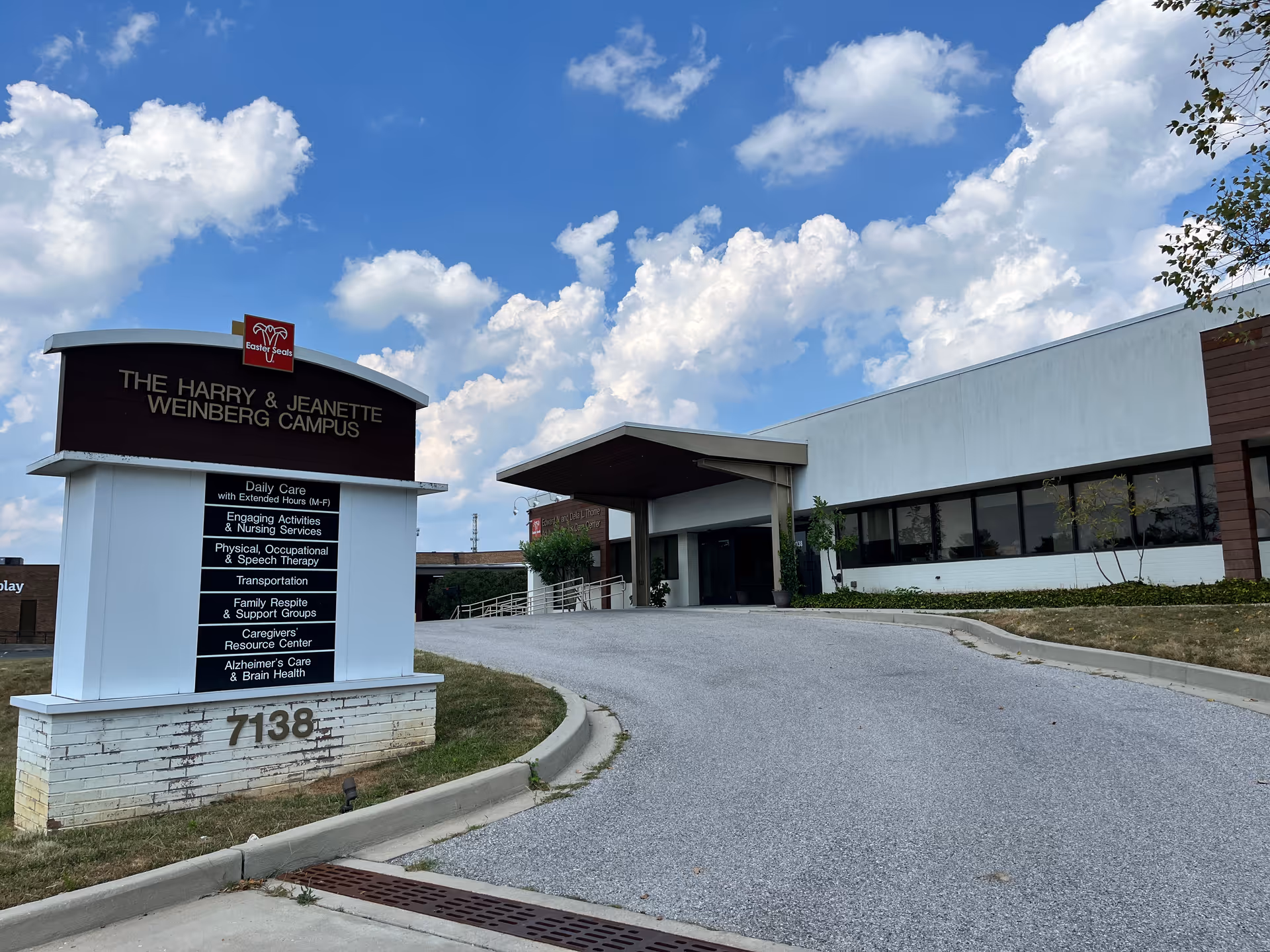 Exterior view of The Harry & Jeanette Weinberg Campus at Easterseals Adult Day Services in Baltimore, showing a large white building with a covered entrance and a sign listing services such as daily care, engaging activities, nursing services, therapy, transportation, family respite, caregiver resource center, and Alzheimer's care. The sky is partly cloudy with blue patches.