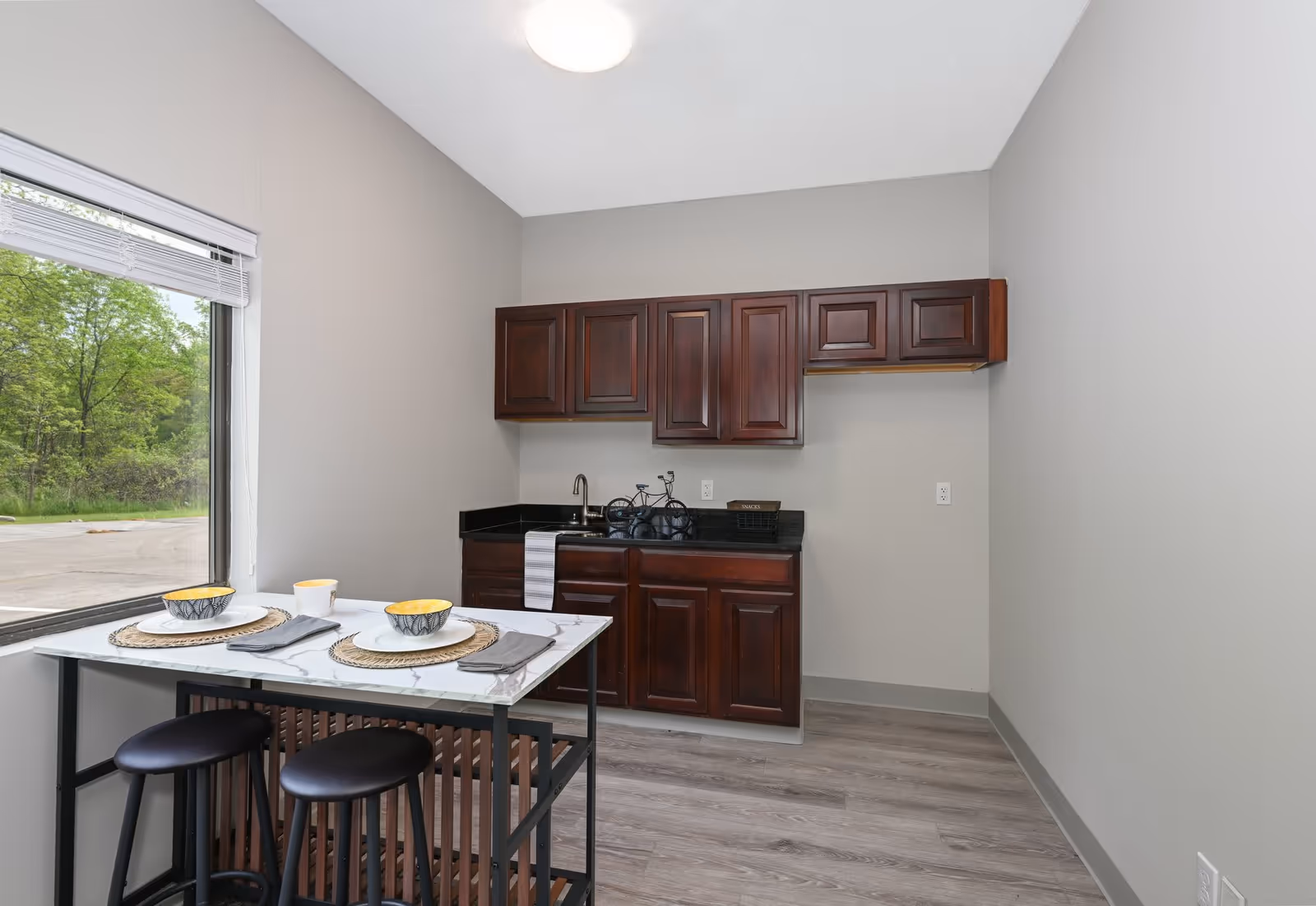 Small kitchen area with dark wooden cabinets, a black countertop, and a sink. In front of the kitchen is a small dining table set for two with bowls, plates, cups, and napkins. A large window next to the table shows green trees outside. The room has light gray walls and wood-look flooring.