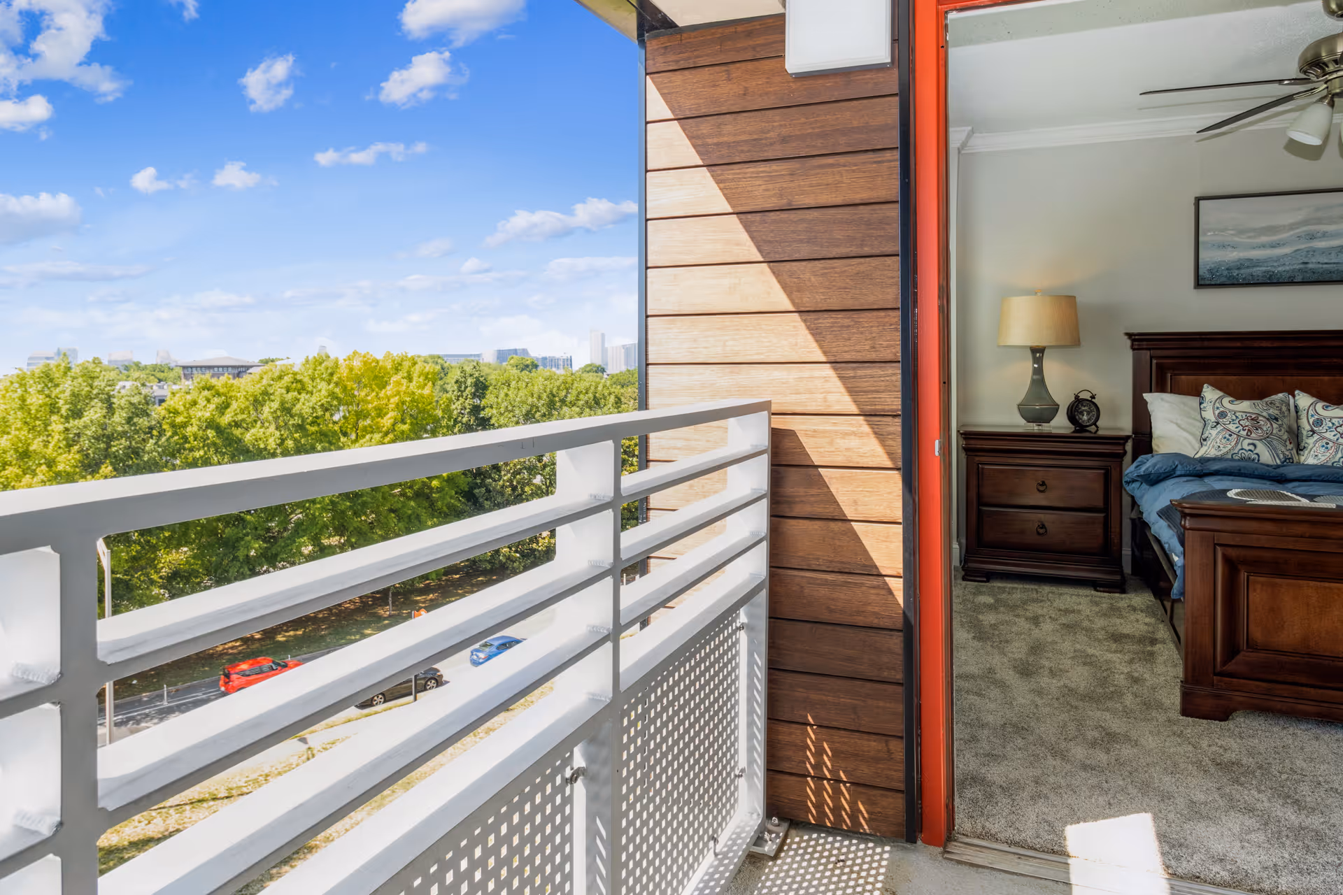 View from a balcony showing a white railing and a street with cars below, with green trees and a blue sky with clouds in the background. To the right, an open door leads into a bedroom with a wooden bed, nightstand with a lamp and clock, carpeted floor, and a ceiling fan.