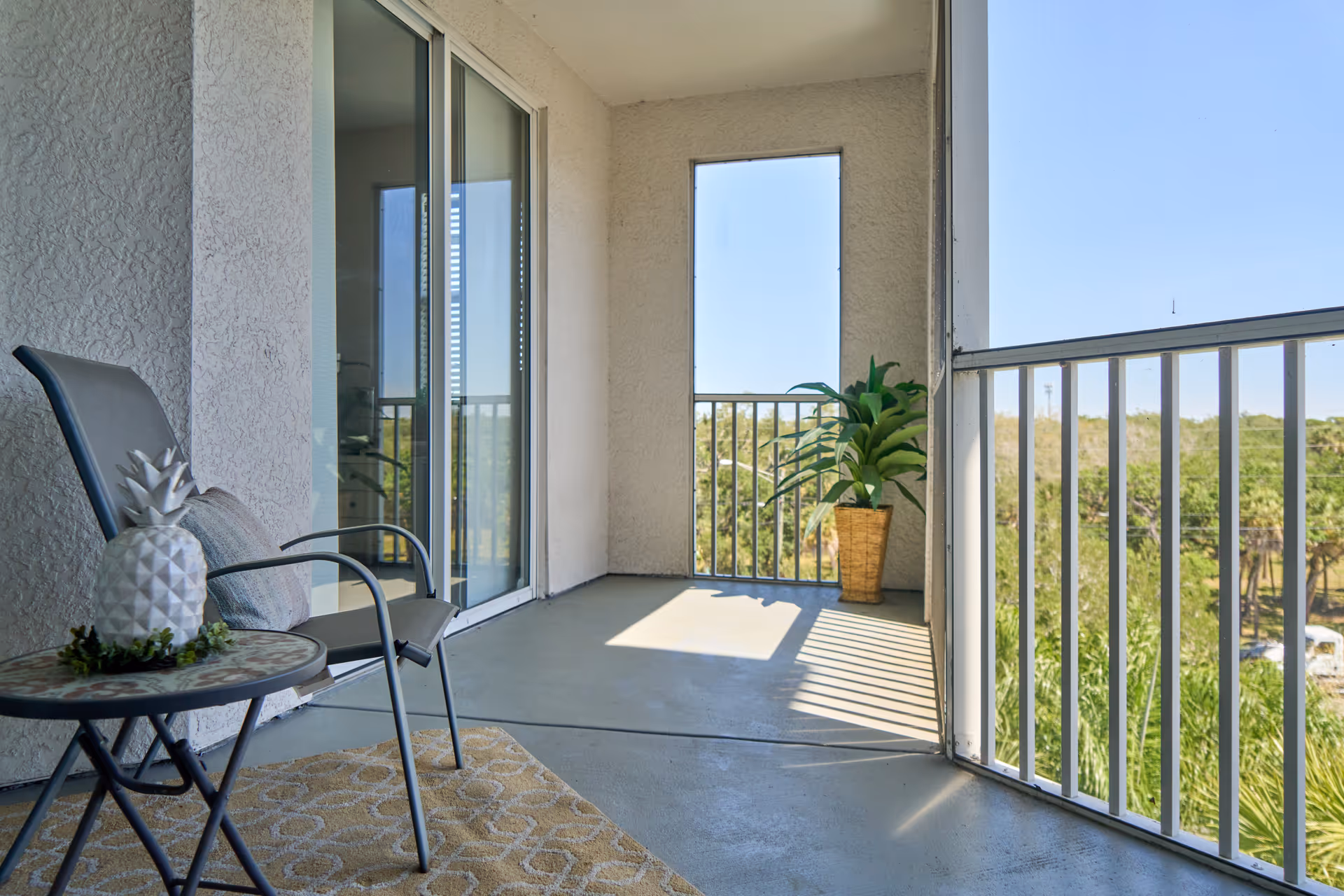 A screened balcony with a metal chair and small round table holding a decorative white pineapple. There is a potted plant in the corner and a sliding glass door leading inside. The balcony overlooks green trees under a clear blue sky.