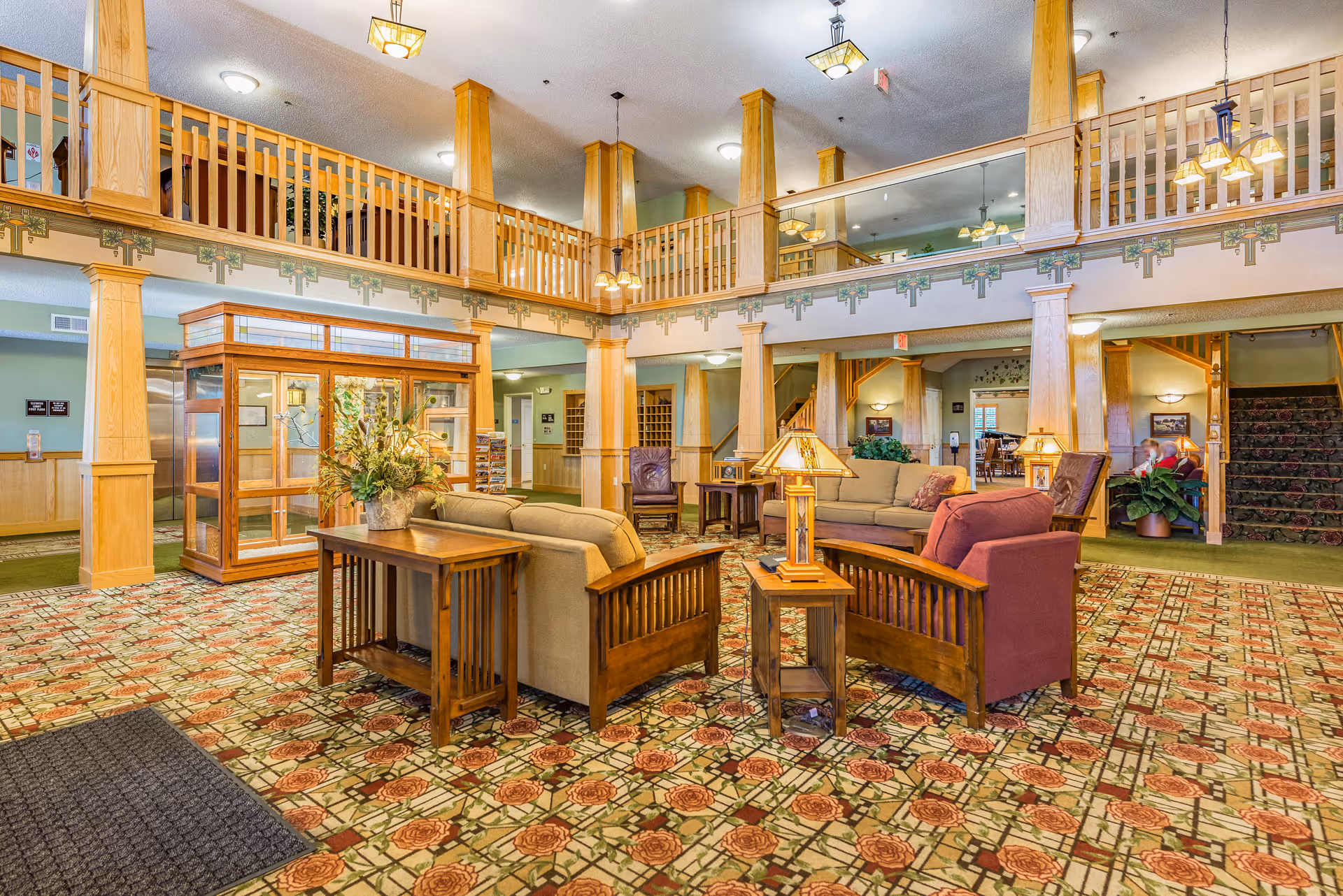 Spacious senior living facility common area with patterned carpet, wooden furniture including sofas, armchairs, side tables with lamps, and a decorative plant centerpiece. The room features wooden columns and railings on the upper level, with warm lighting and a welcoming atmosphere.