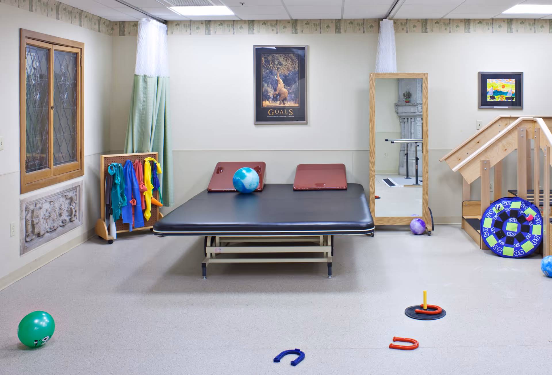 A therapy or exercise room with a large black padded table in the center, a blue and white ball on the table, colorful exercise bands hanging on a rack to the left, a full-length mirror on the right wall, and various therapy equipment including a target board and small balls scattered on the floor.