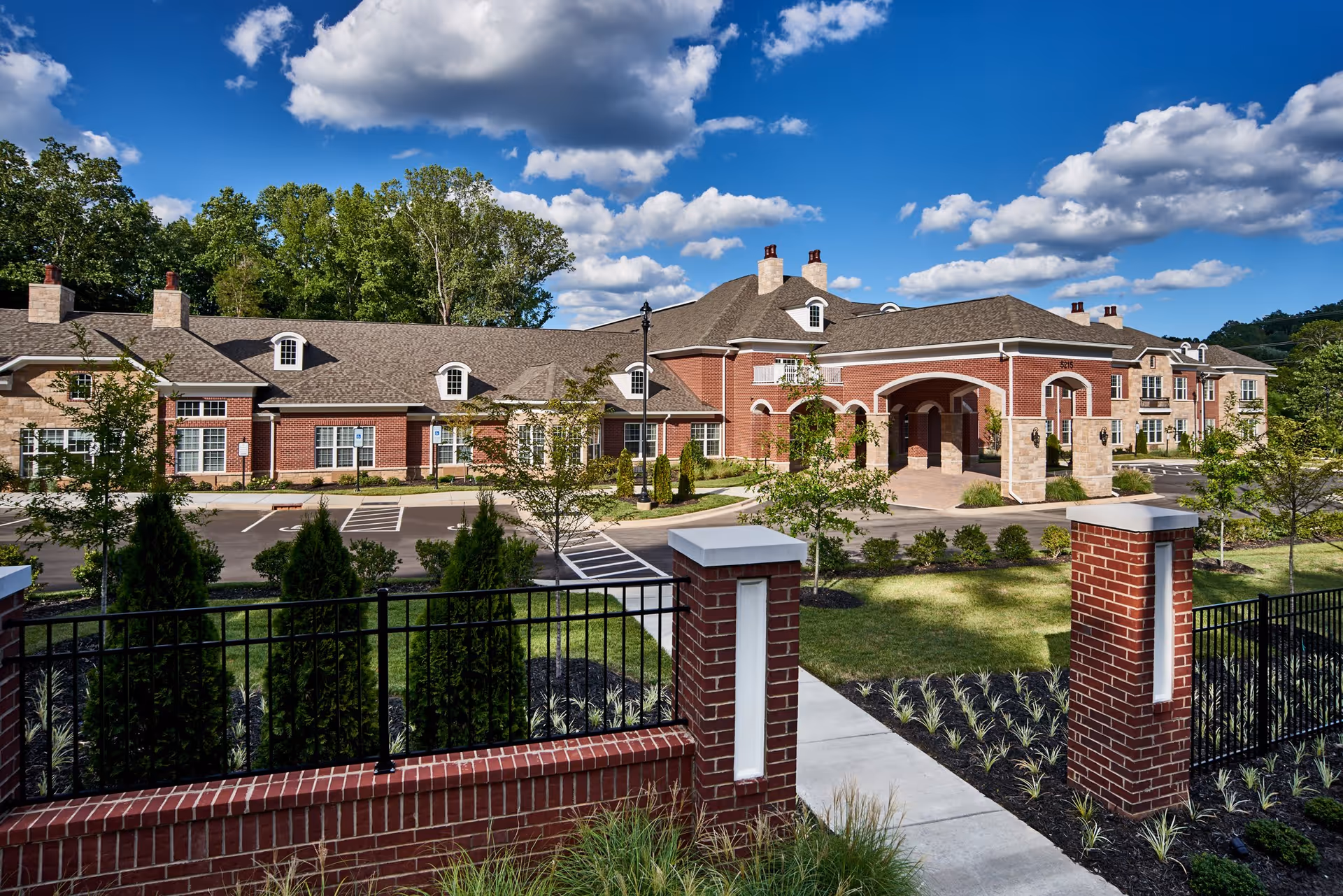 Exterior view of a large brick senior living facility building with multiple chimneys, arched entrance, and a well-maintained landscaped garden and parking area under a blue sky with scattered clouds.