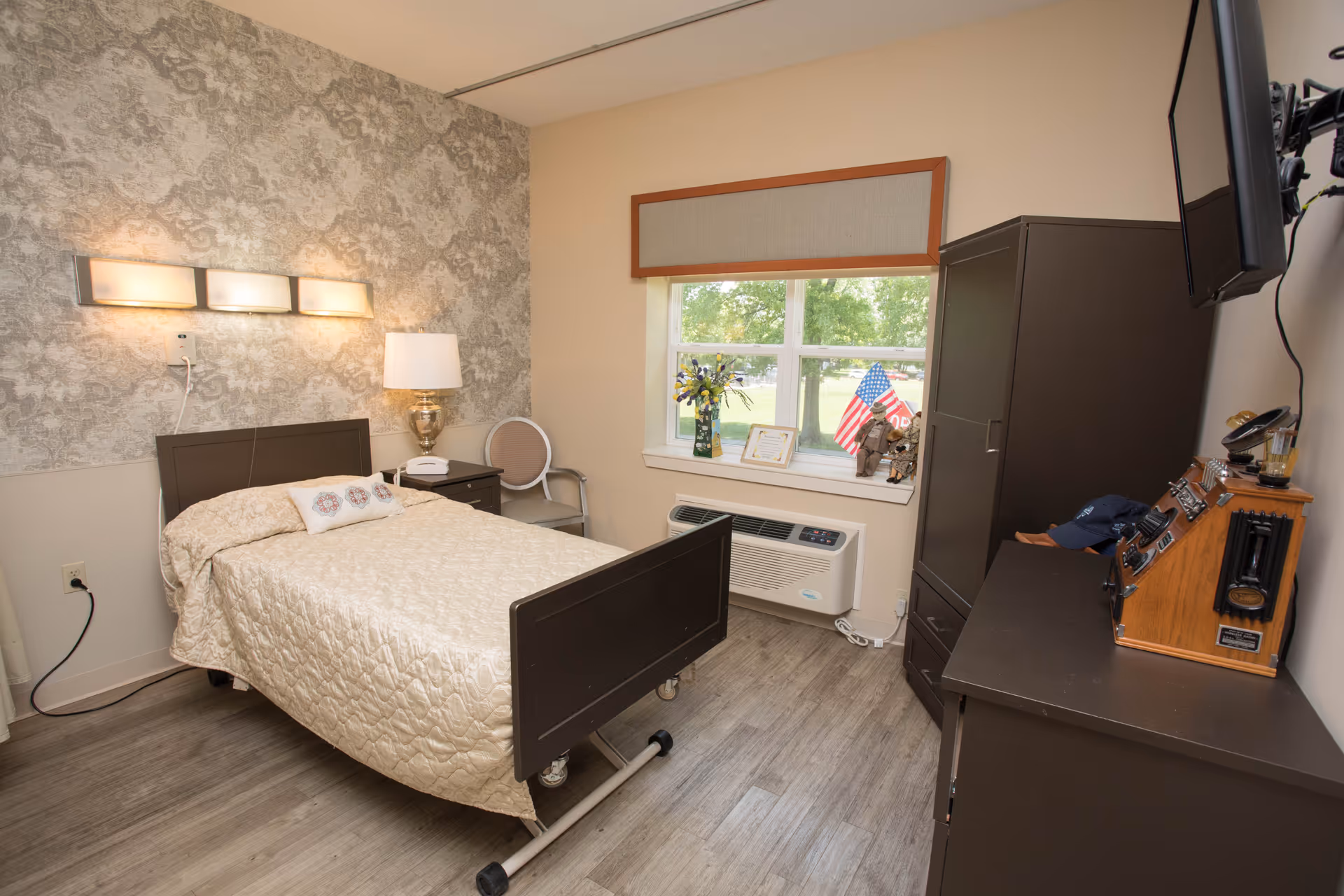 A neatly arranged bedroom in a senior living facility with a single bed covered in a beige quilt, a bedside table with a lamp, a chair, a window with a view of greenery outside, and a dresser with a mounted TV above it. The room has light-colored walls, wood flooring, and decorative wallpaper on one wall.