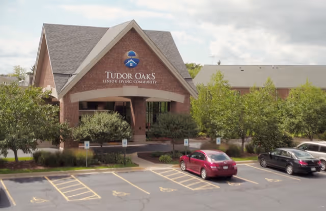 Front exterior view of Tudor Oaks Senior Living Community building with a brick facade, a peaked roof, and a covered entrance. Several cars are parked in the parking lot in front, with trees and shrubs surrounding the entrance area.