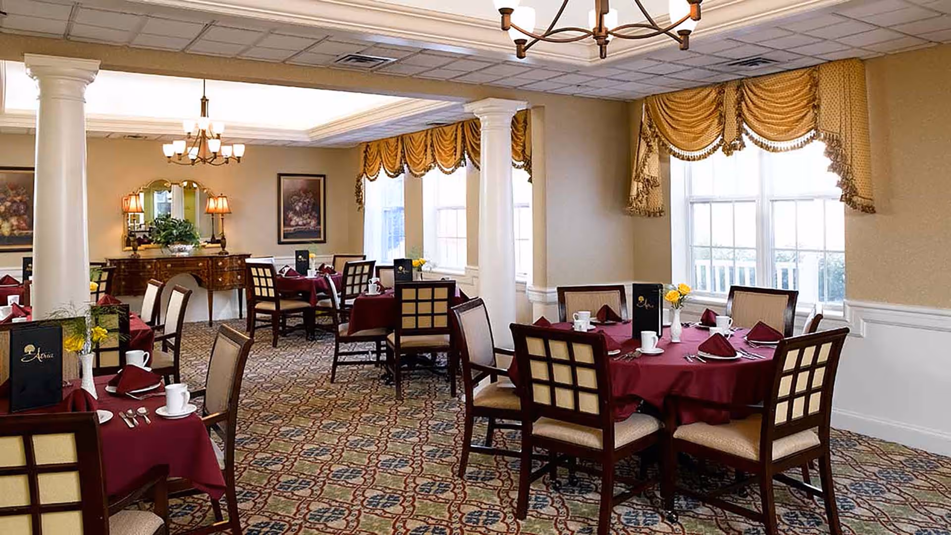 Formal dining room with round tables set with burgundy tablecloths and place settings, chandeliers, and draped windows.