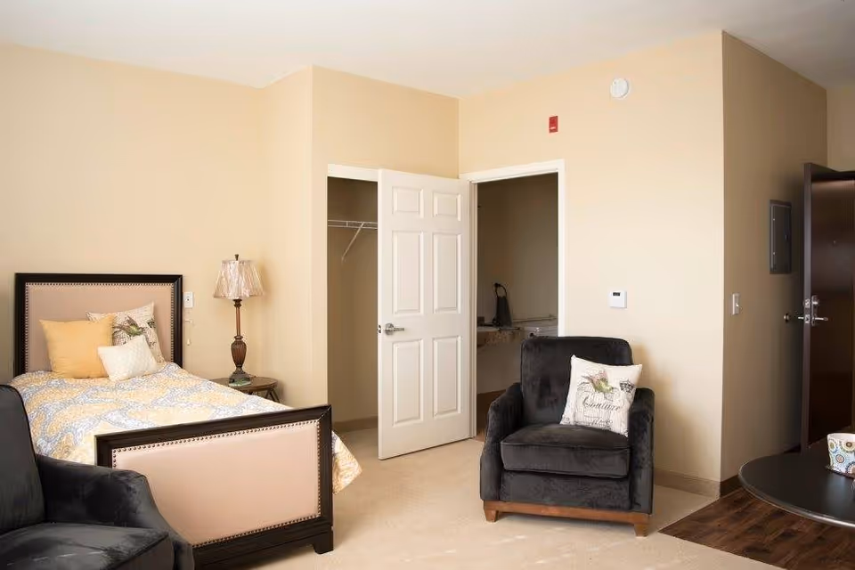 A cozy senior living bedroom with a single bed featuring a beige and yellow patterned comforter and multiple pillows. There is a dark upholstered armchair with a decorative pillow, a small side table with a lamp, an open closet door, and an open bathroom door. The walls are painted beige and the floor is carpeted.