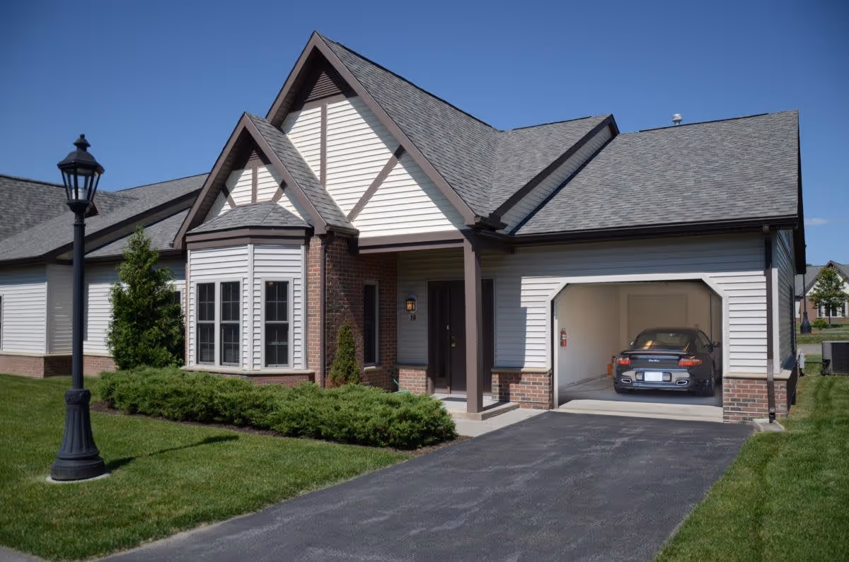 Exterior view of a single-story residential building with a gabled roof, white siding, and brick accents. There is a driveway leading to an open garage with a car parked inside. A lamp post and well-maintained green shrubs and lawn are visible in the front yard under a clear blue sky.