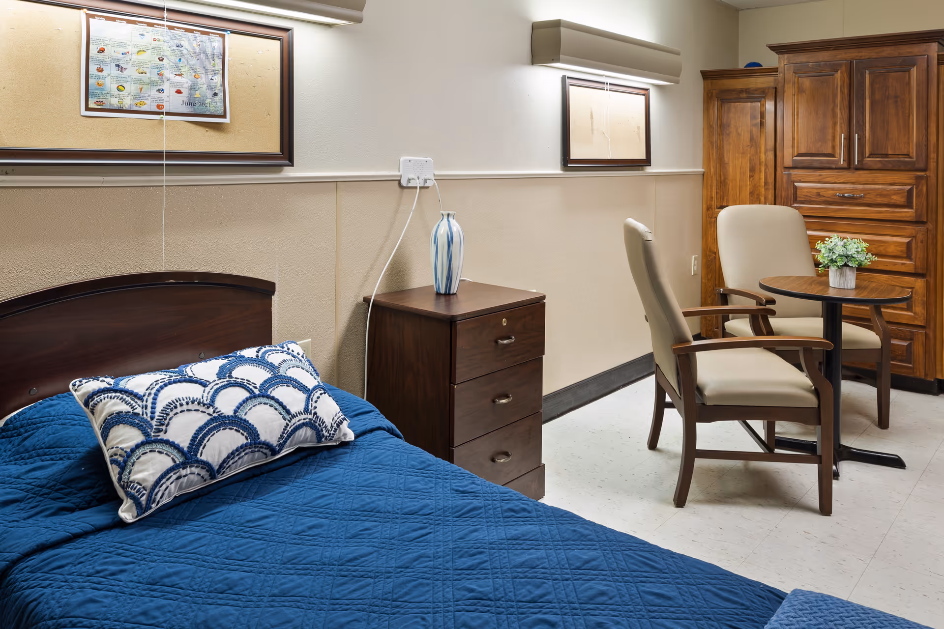 A senior living facility bedroom with a bed covered in a blue quilt and a decorative pillow. Next to the bed is a wooden nightstand with a blue and white vase on top. On the wall above the nightstand is a bulletin board with a calendar pinned to it. Across from the bed, there is a small round table with two beige cushioned chairs and a small potted plant. In the background, there is a large wooden wardrobe.