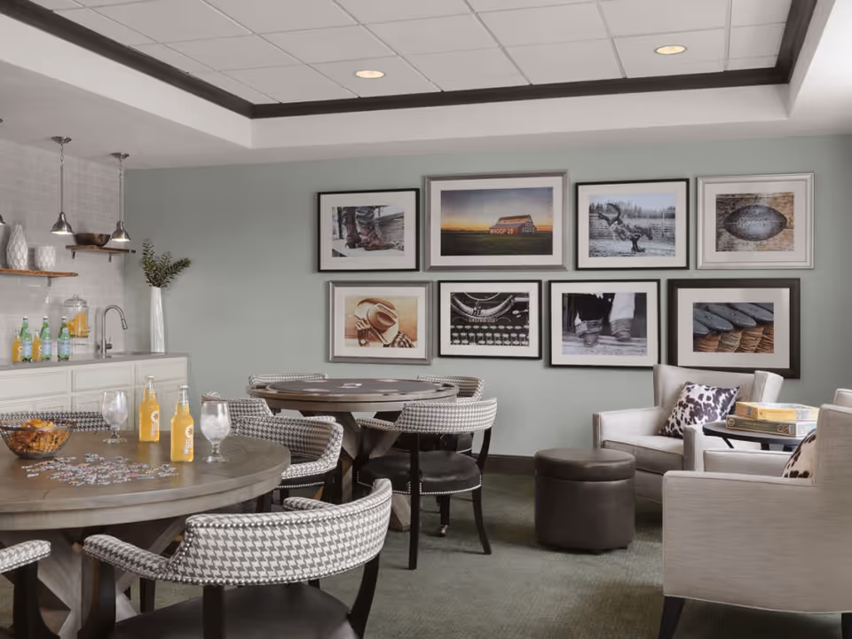 A cozy common area in a senior living facility with round tables and cushioned chairs arranged for socializing or activities. The walls are decorated with framed pictures, and there is a small counter with a sink, beverages, and decorative items. Two armchairs with patterned pillows and a small table with board games are also visible.