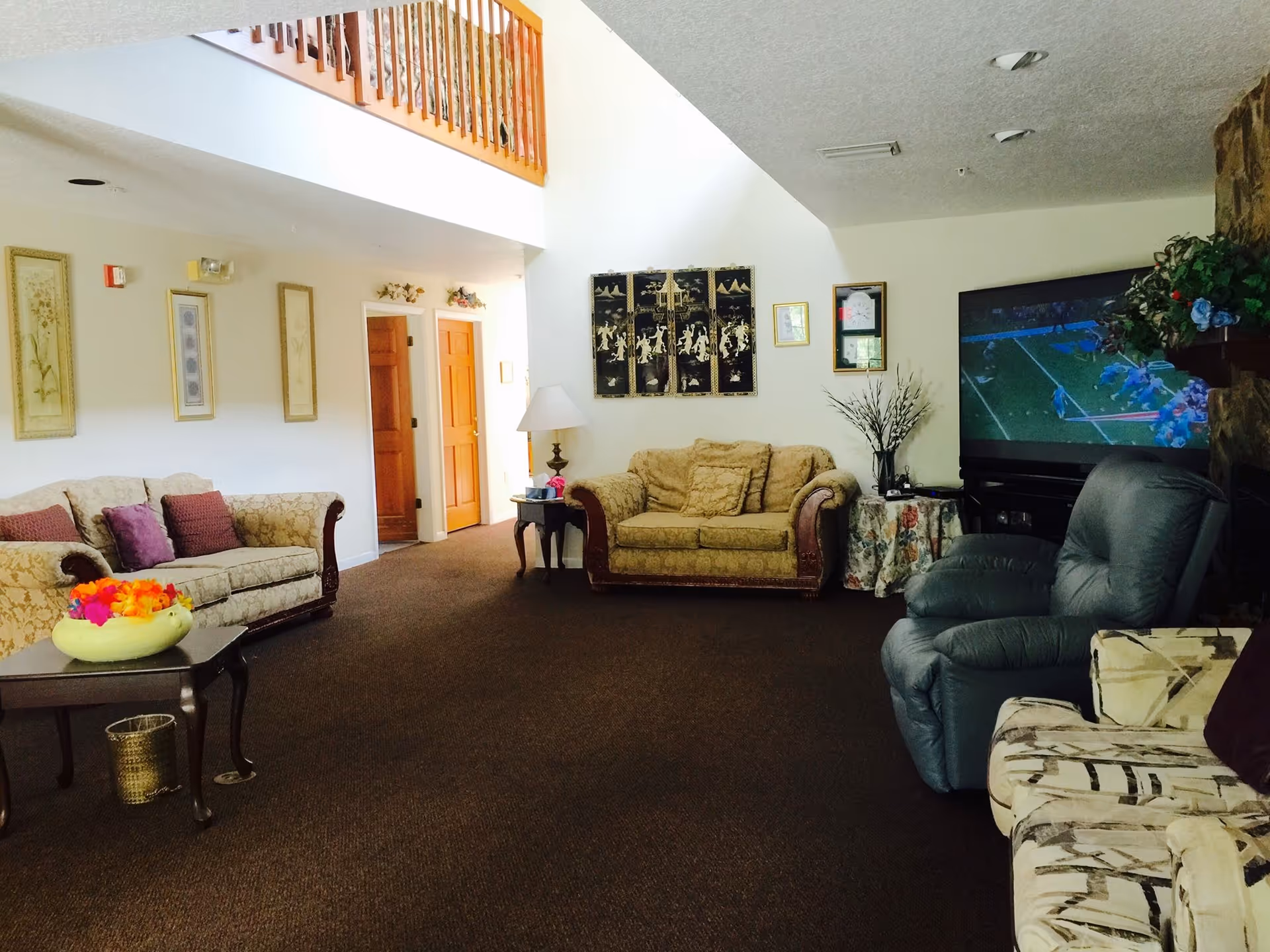 A spacious living room with multiple sofas and armchairs arranged around a large flat-screen TV showing a football game. The room features a high ceiling with a wooden railing balcony above, framed artwork on the walls, a side table with a lamp, and decorative plants.